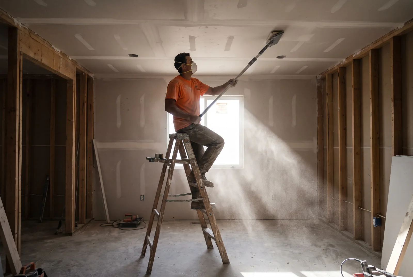 A worker sanding a drywall ceiling, dust caught in a shaft of daylight