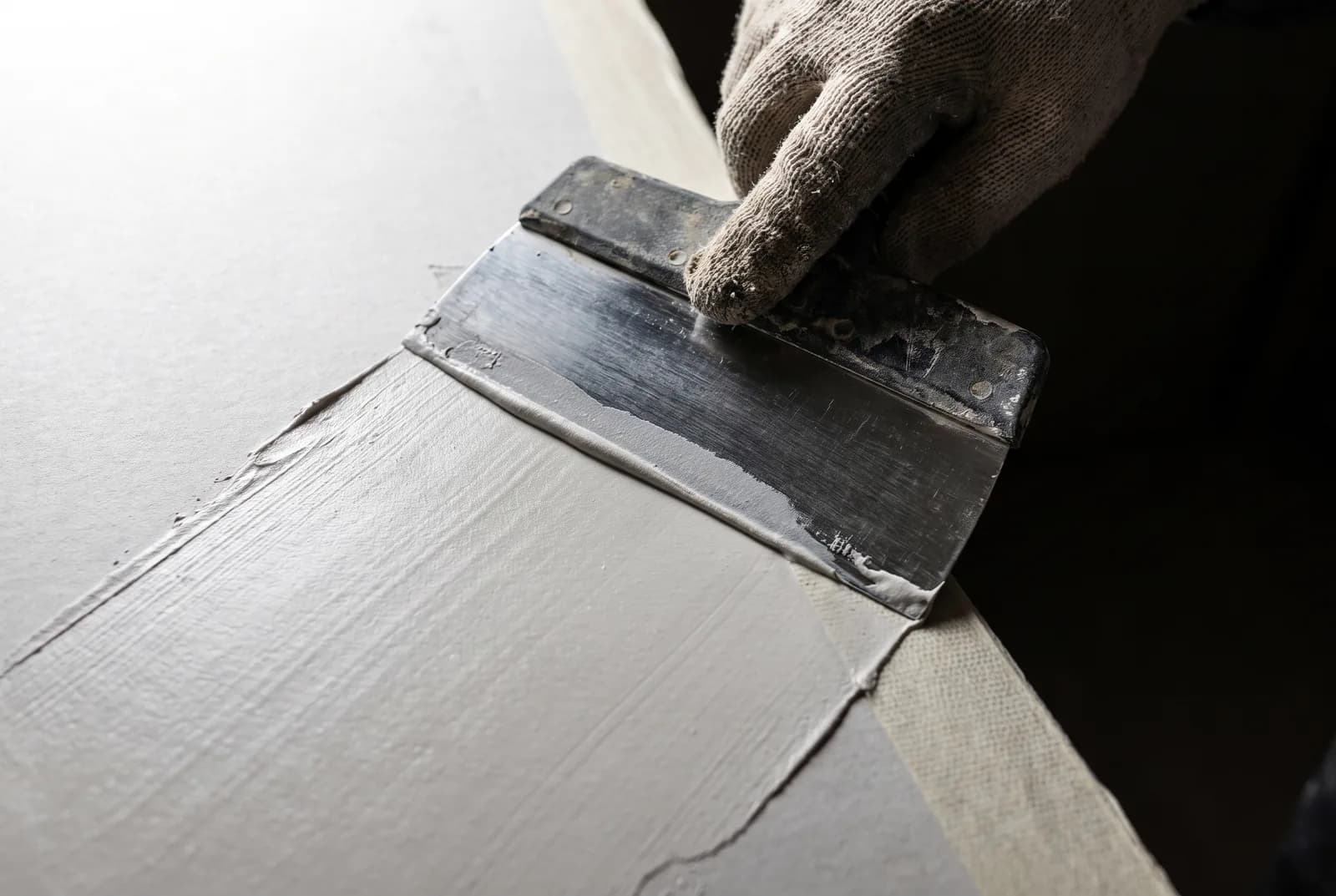 A taping knife drawing joint compound across a drywall seam under raking light